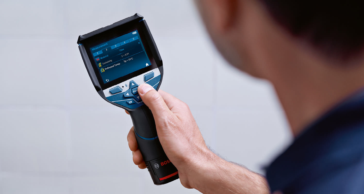 Käsi pitelee sinimustaa Bosch-lämpökameraa valkoista laattaseinää kohti