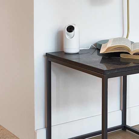 Tummapintainen sivupöytä, valkoinen valvontakamera, avonainen kirja ja kultainen lampunjalka