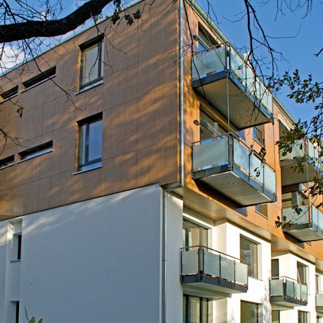 Moderni kerrostalo: vaalea alaosa, puupaneelit yläkerrassa, lasikaiteiset parvekkeet, sinitaivas