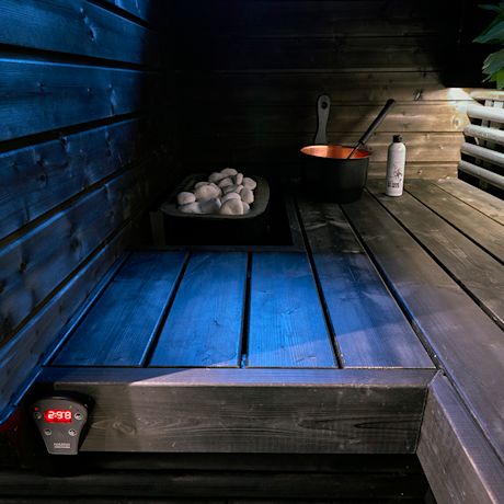 Sinisellä valaistulla puusaunan lauteella näkyy kiuas kivineen, kuparikiulu, kauha ja pullo.