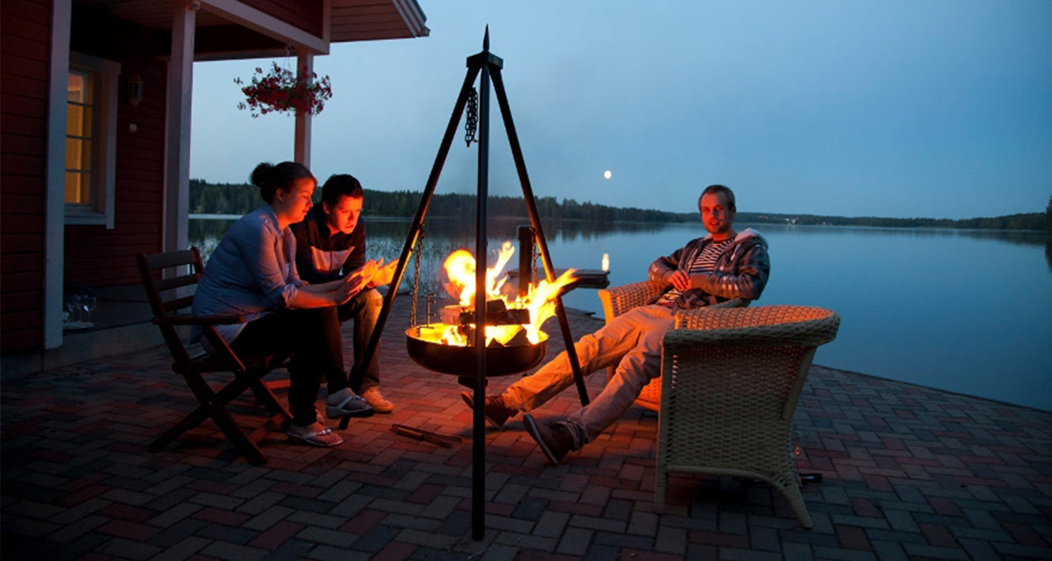 Kolme ihmistä istuu avotulen ympärillä laituriterassilla järven rannassa hämärässä illassa.