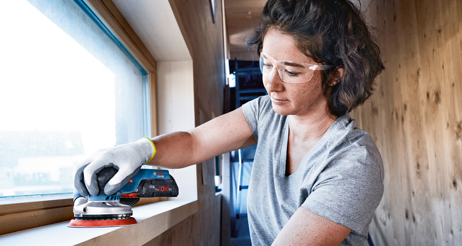 Henkilö hioo sinimustalla ProCORE 18V -sähköhiomakoneella vaaleaa puista ikkunankarmia.