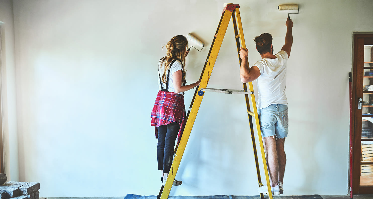 Mies ja nainen maalaavat rullilla valkoista sisäseinää keltaisilta tikkailta.