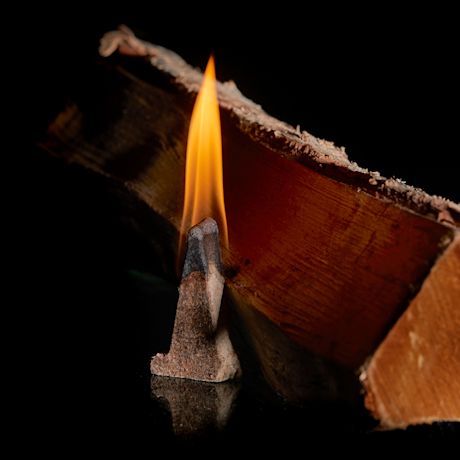 Oranssi liekki palaa ruskealla suitsukekartiolla mustan pinnan yllä, taustalla puunriekale.