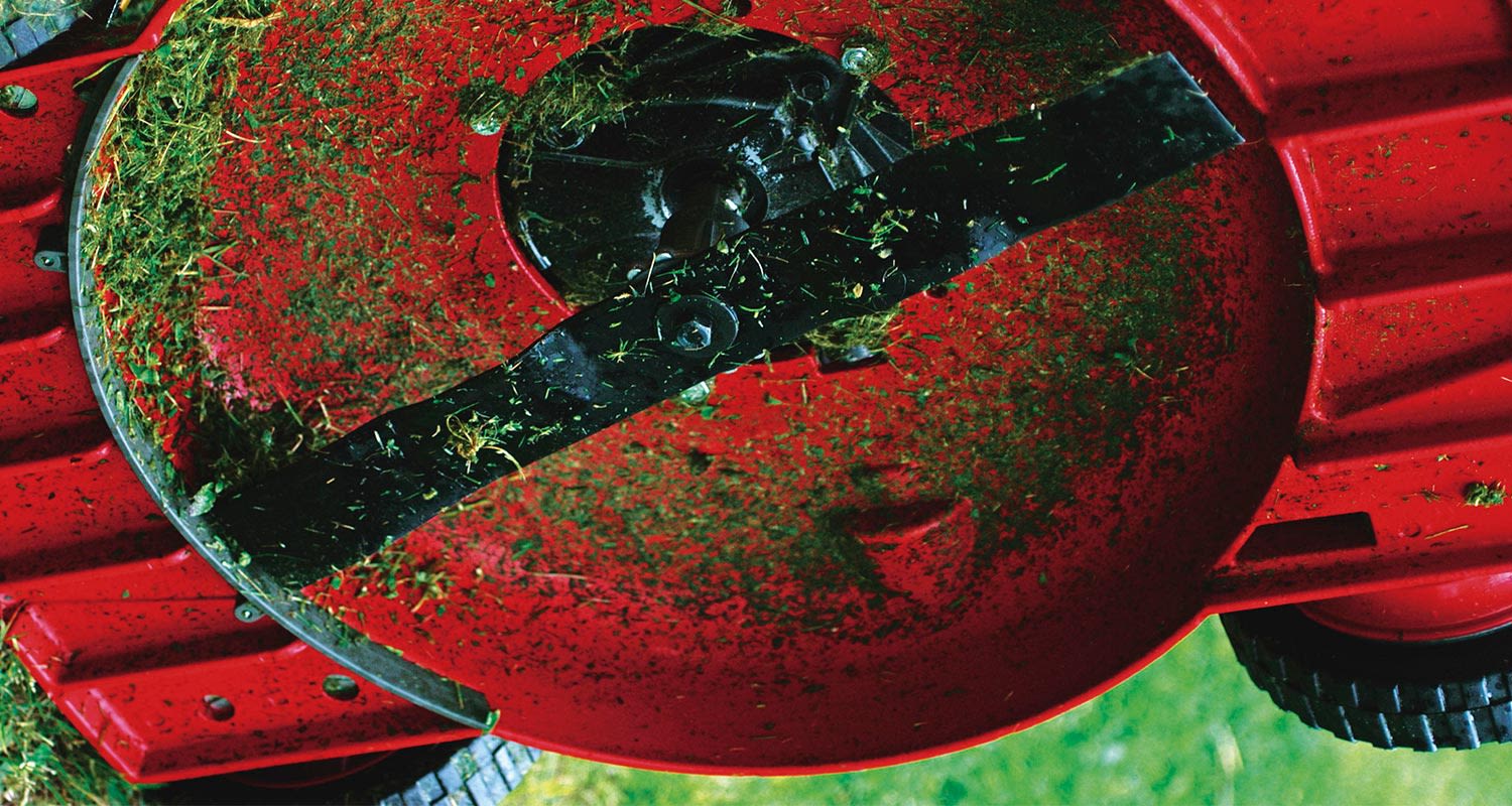 Punaisen ruohonleikkurin alaspäin käännetty kansi, mustat terät ja vihreät ruohojäämät.