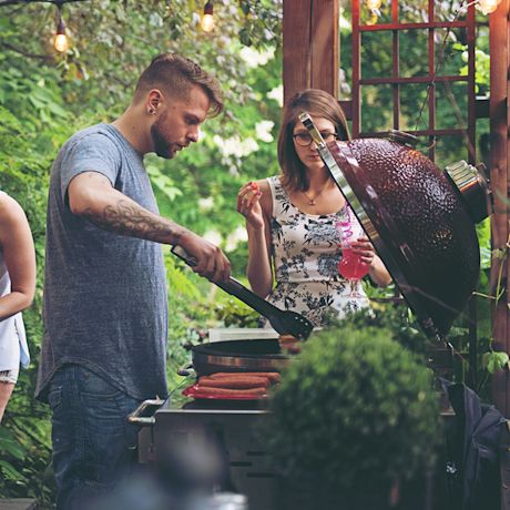 Ulkogrillillä t-paidallinen mies kääntelee makkaraa, nainen pitää mansikkaa ja pinkkiä juomaa.