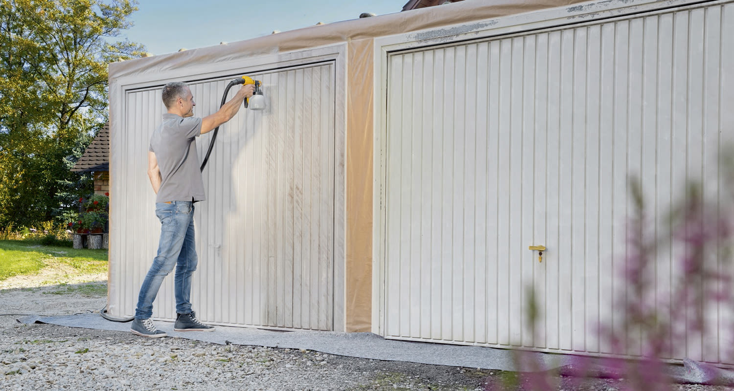 Mies suihkumaalaa valkoiseksi kaksiosaisen autotallinoven pihapiirissä, vihreää taustalla.