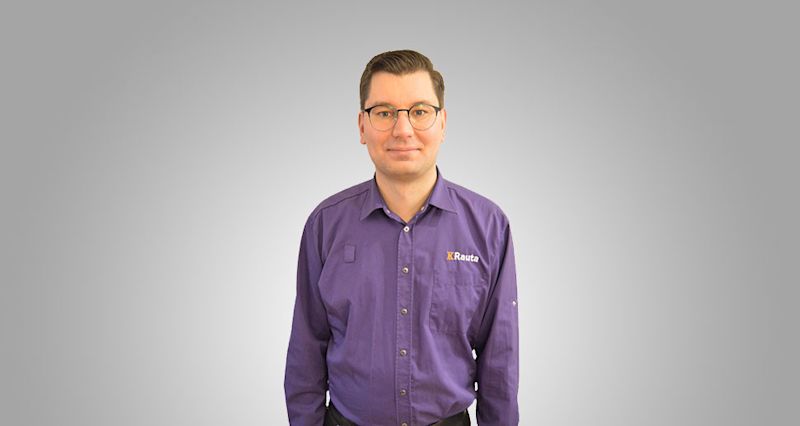 Mies seisoo harmaalla taustalla, yllään violetti K-Rauta-työasu.