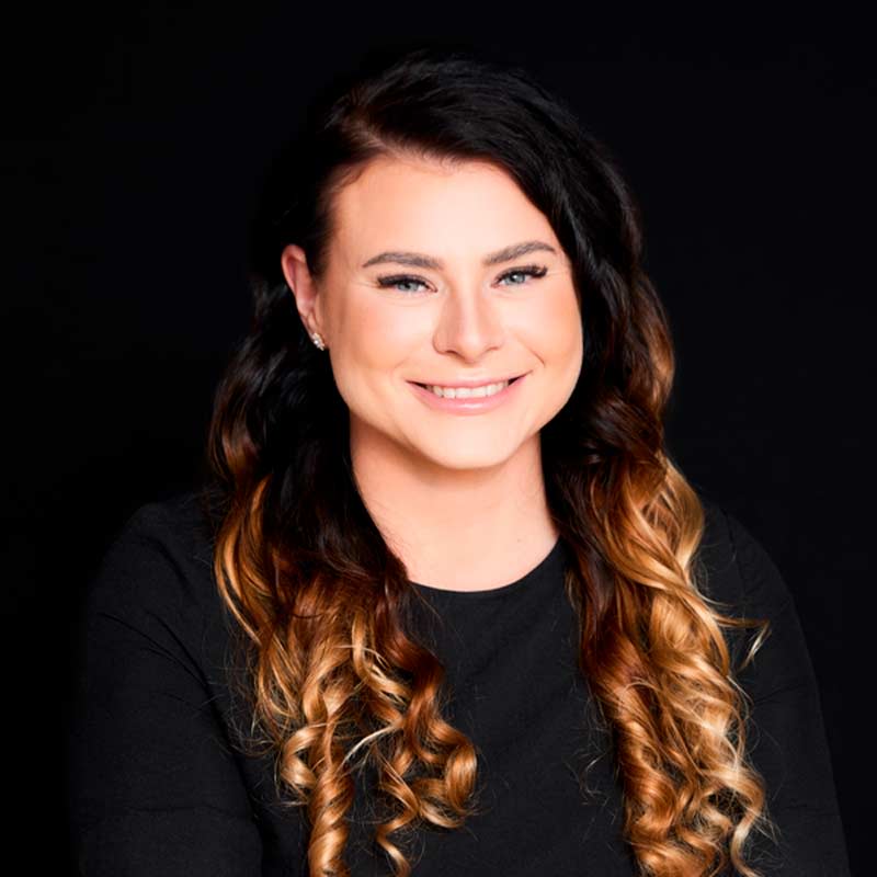 Henkilö mustassa paidassa, pitkät ruskea-vaaleat kiharat tummalla taustalla.