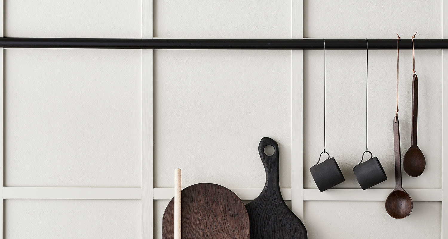 Valkoisella paneeliseinällä musta tanko, roikkuvat mustat mukit, puulusikat ja ruskeat leikkuulaudat.