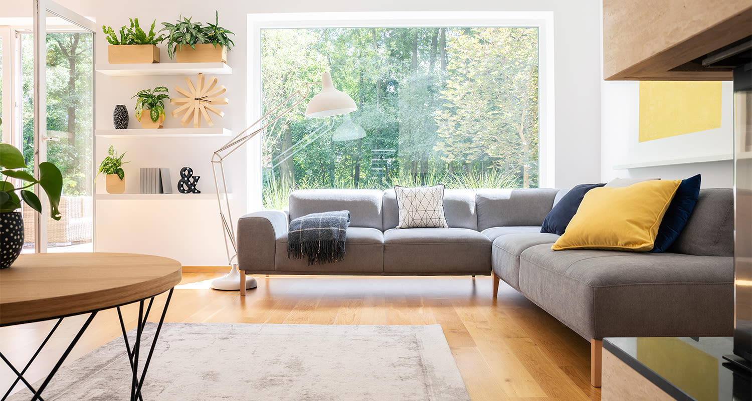 Valoisa olohuone: harmaa kulmasohva keltatyynyllä, iso ikkuna metsämaisemaan, puupöytä ja kasvit.