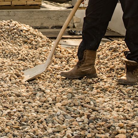 Henkilö ruskeissa työkengissä sorakasassa lapion varressa rakennustyömaalla.