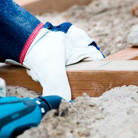 Valko-sinipunainen työkäsine pitelee puulautaa hiekalla, edessä sinimusta akkuporakone