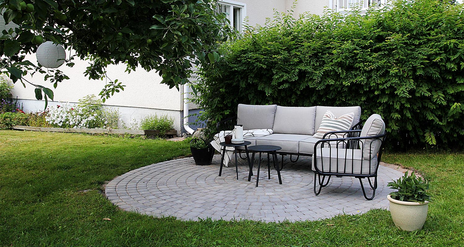 Vihreässä puutarhassa harmaa sohva ja nojatuoli mustilla metallijaloilla pyöreällä kiviterassilla.