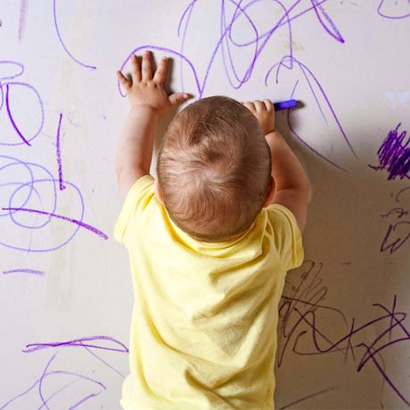 Taapero keltaisessa paidassa piirtää violettikynällä valkoiselle seinälle.