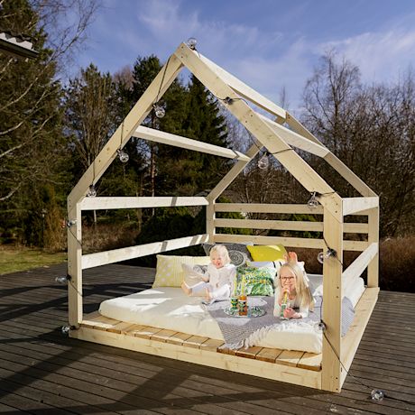 Puinen mökkikehikko terassilla, valosarja, patja, värikkäät tyynyt ja lapset keväisessä pihalla.