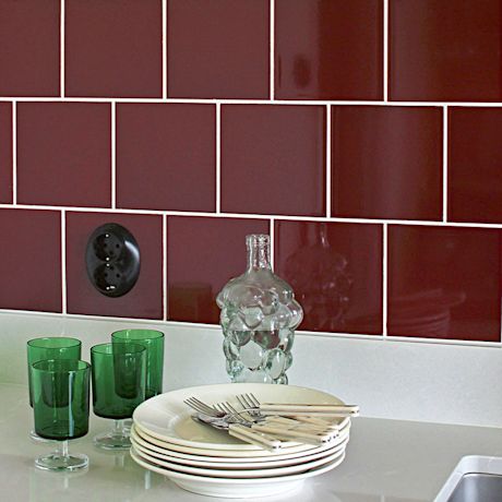 Valkoiset lautaset pinossa aterimineen, vihreitä laseja ja lasipullo tummanpunaista laattaa vasten