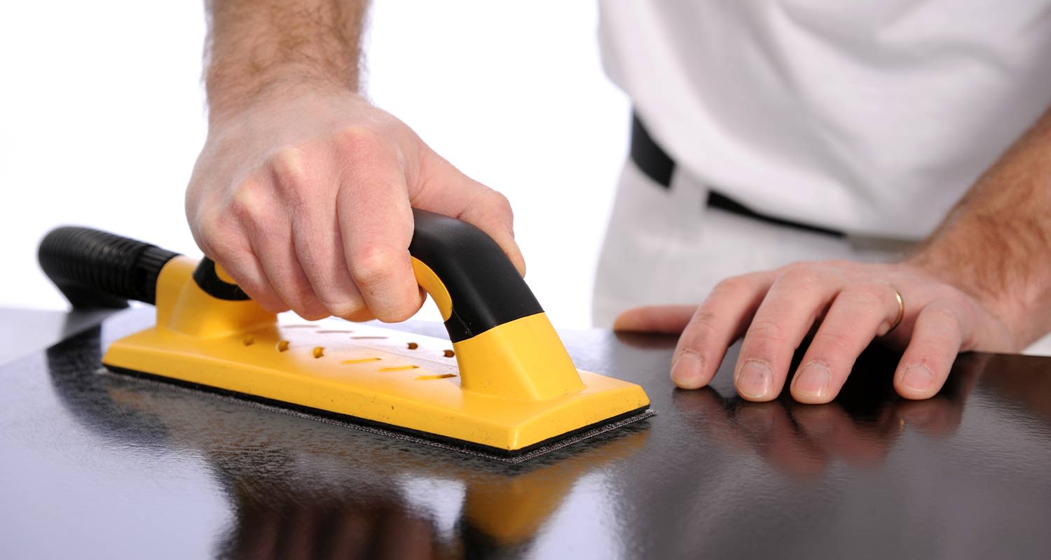 Henkilö hioo mustaa kiiltävää pintaa kelta-mustalla käsihiomatyökalulla valkoisessa tilassa.