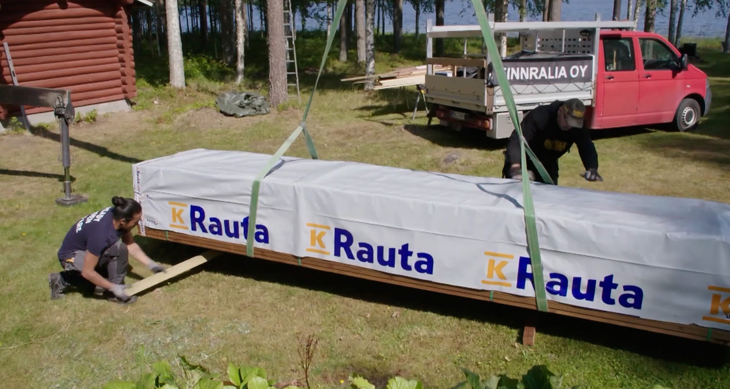 K-Rauta-pakkaus nostetaan nurmella, taustalla punainen pakettiauto ja metsä