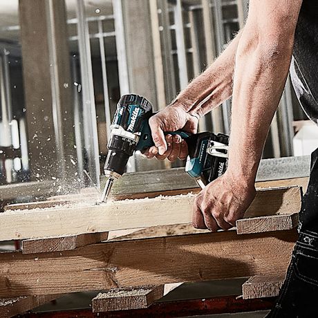 Rakennustyömaalla turkoosi-musta Makita-porakone poraa puupalkkia, puusilppu lentää.