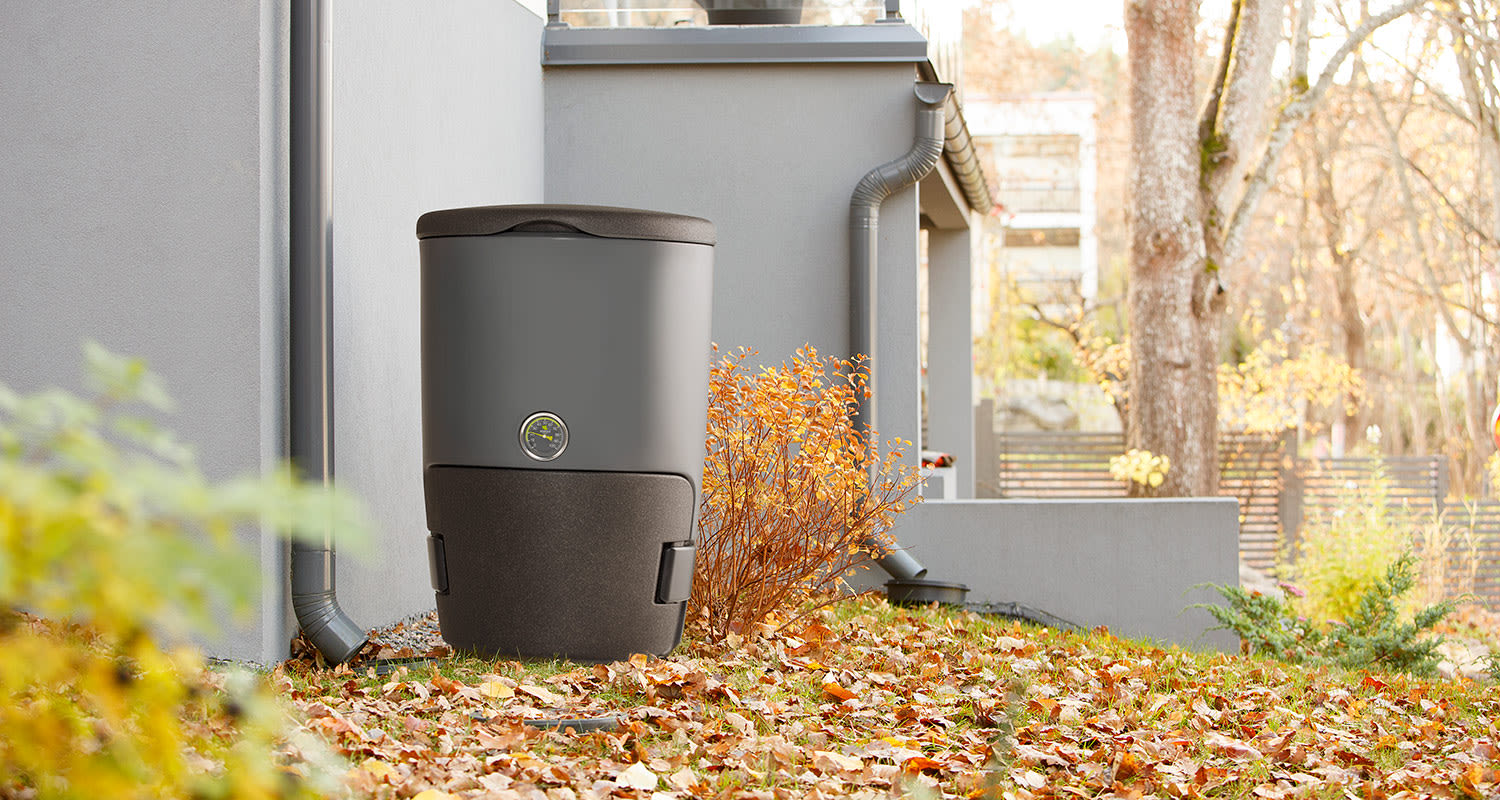 Harmaa sadevesisäiliö talon seinustalla, syksyiset lehdet ja oranssit pensaat.