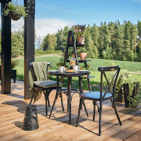 Puupatio maalaismaisemassa, musta pöytä kahdella tuolilla, kahvikupit, leivokset ja ruukkukukat.