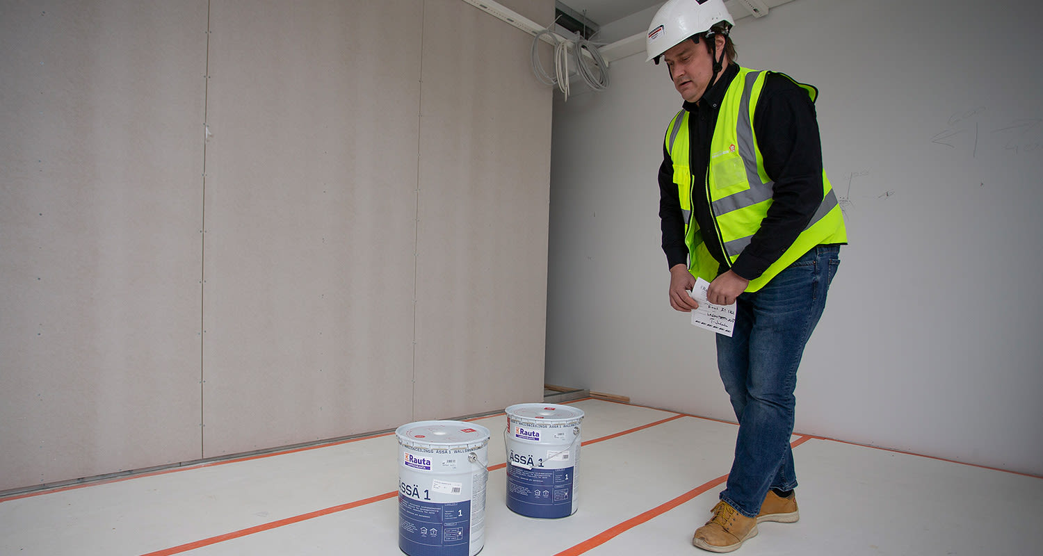 Keltaliivinen kypärätyöntekijä tarkastelee kahta valko-sinistä Rauta-maalipurkkia tyhjässä huoneessa