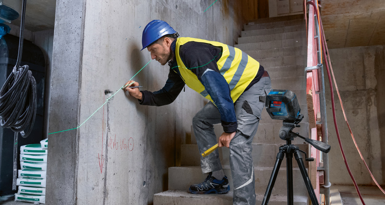 Keltaliivinen työmies sinisessä kypärässä merkitsee betoniseinää vihreällä lasersäteellä.