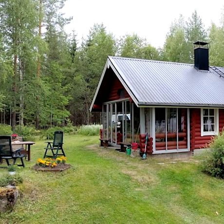 Punainen hirsimökki metallikatolla ja lasitetulla kuistilla metsäaukealla, pihatuolit nurmikolla