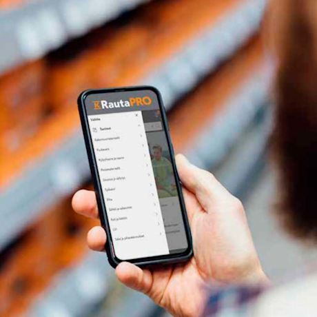 Henkilö pitelee puhelinta, näytöllä RautaPRO-sovelluksen valikko, oranssit hyllyt taustalla.