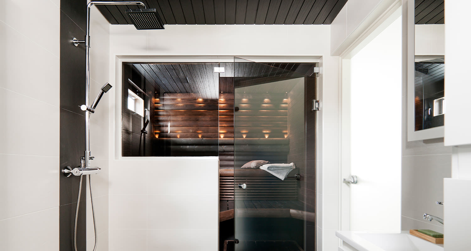 Moderni valkoinen kylpyhuone suihkulla ja lasiseinällä, takana puusauna lämpimillä valoilla.