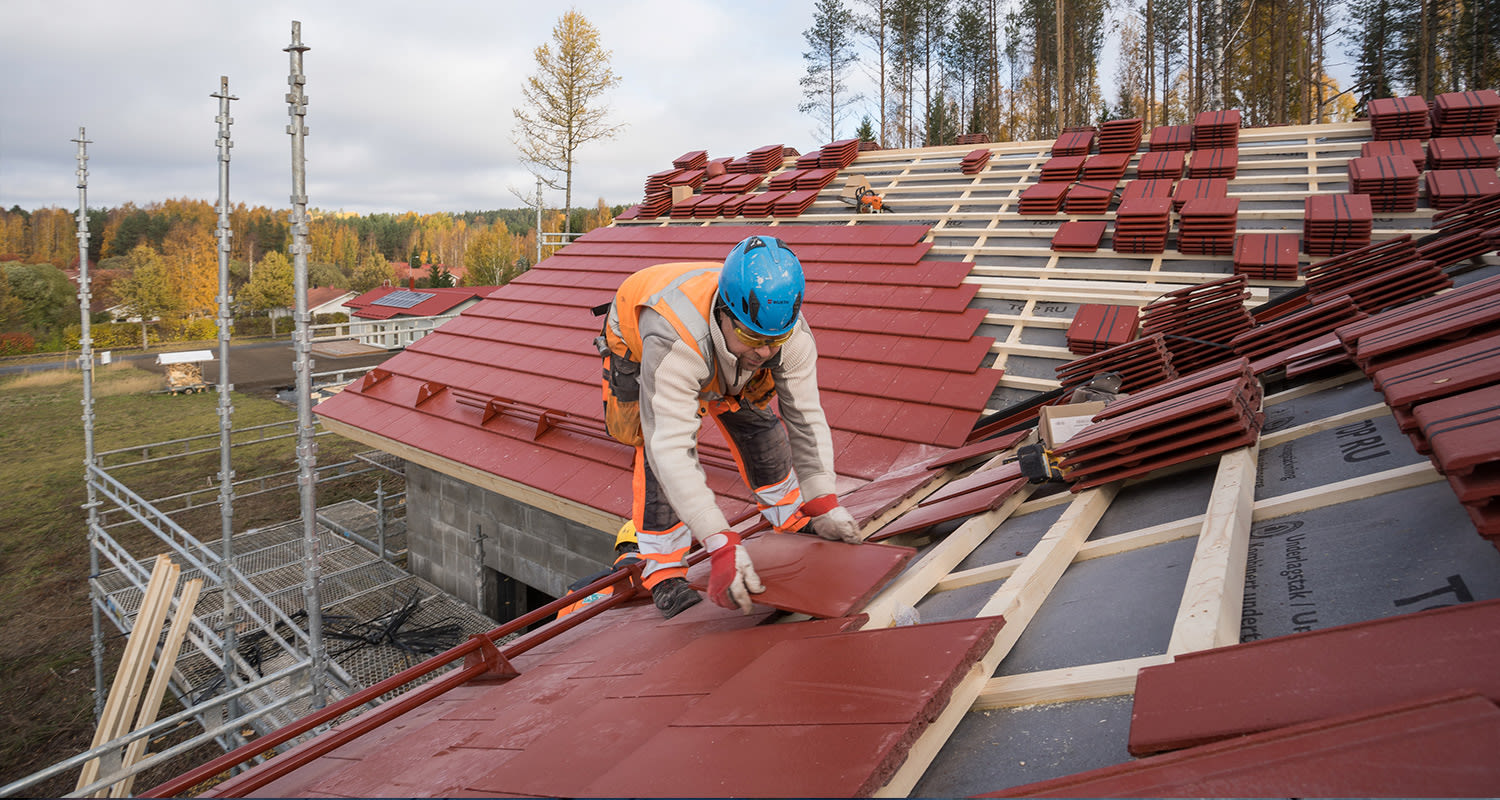 Rakennustyömies asentaa punaisia kattotiiliä puurunkoiselle katolle metsän reunassa.