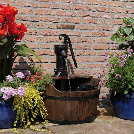 Puinen tynnyri mustalla käsipumpulla, vesivirta, punaiset ja violetit kukat tiiliseinää vasten