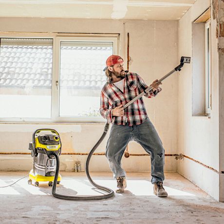 Remonttitilassa ruutupaitainen mies soittaa imurin letkulla kuin kitaralla, keltainen imuri.