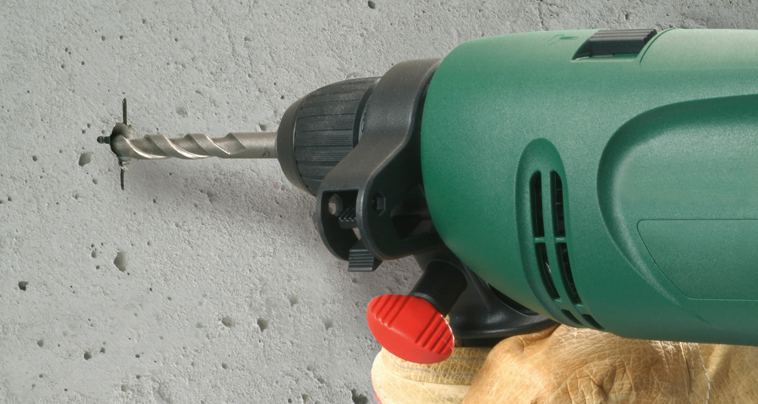 Vihreä sähköpora punaisella vivulla ja suojakäsine poraa harmaata betoniseinää