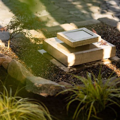 Harmaat betonilaatat vesiaiheena pikkupuutarhassa, ympärillä purukiveä, kasveja ja auringonvalo.