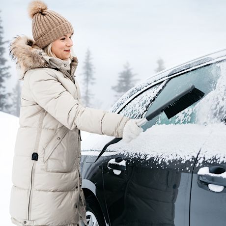Henkilö beigen talvitakissa ja pipossa pyyhkii lumet mustan auton tuulilasista lumisessa metsässä