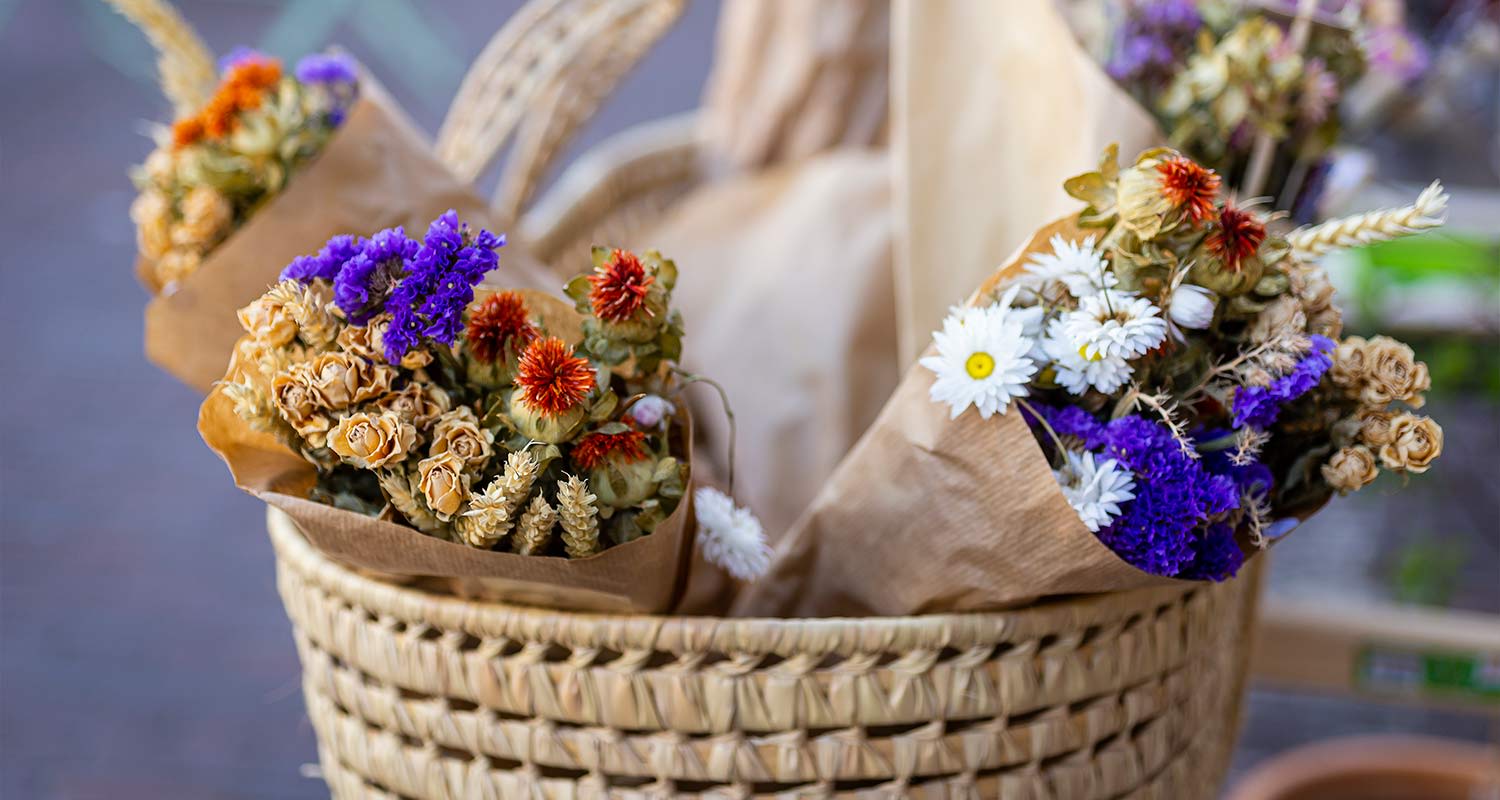 Kuivakukkakimput ruskeassa paperissa rottinkikorissa, violetit, valkoiset ja oranssit kukat.