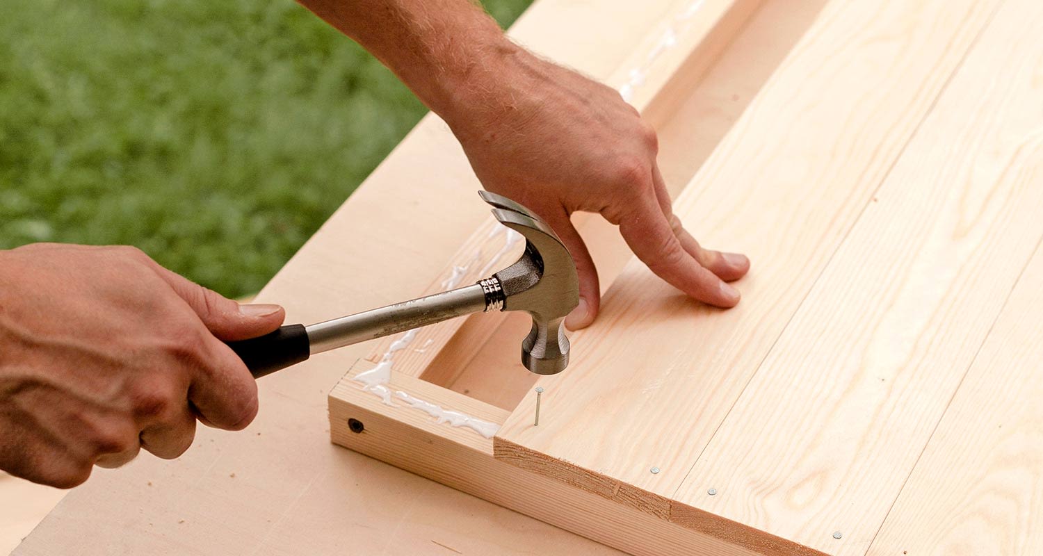 Kädet iskevät vasaralla naulan vaaleaan puulevykehykseen ulkona nurmikolla.