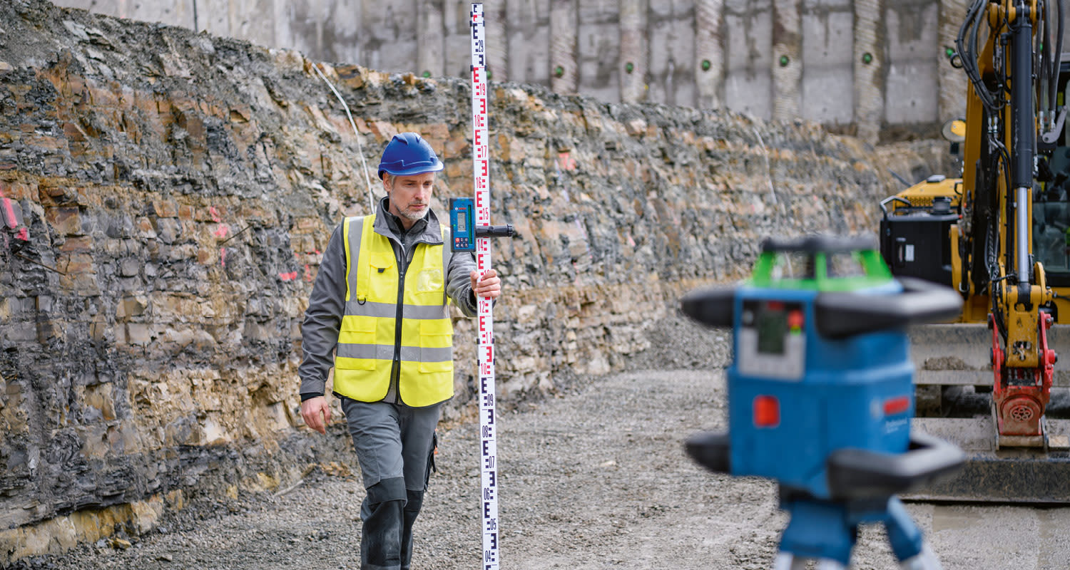 Kaivutyömaalla kypäräpäinen keltaliivinen mittamies pitää mittakeppiä, etualalla sininen laserlaite