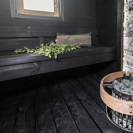 Tumma sauna, penkillä vihreä vihta ja harmaa tyyny, ikkunasta valoa, kiuas täynnä kiviä