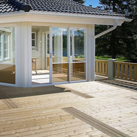 Valkoinen mökki lasiliukuovilla ja vaalea terassi, taustalla metsäinen järvimaisema.