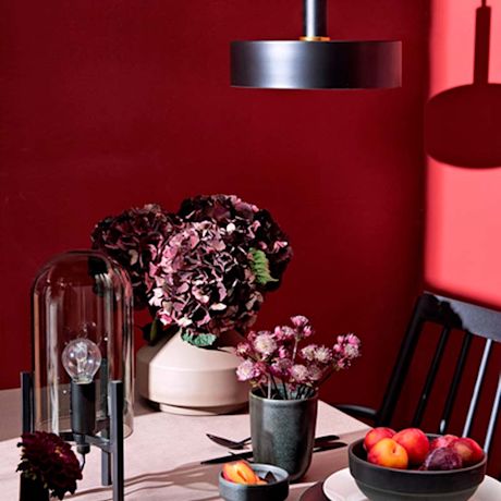 Punaisella seinällä varjostava musta lamppu. Pöydällä pinkit kukkamaljakot ja hedelmäkulho.