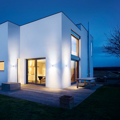 Moderni valkoinen talo iltahämärässä, lämminsävyiset valot, puisella terassilla.