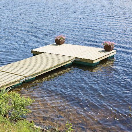 Puisella laiturilla kaksi ruukkua vaaleanpunaisia kukkia, sininen järvi ja vihreä ranta.