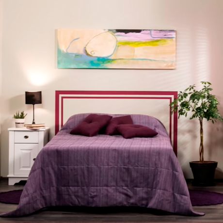 Violettipeitteinen sänky purppuratyynyillä, valkoinen yöpöytä lampulla, viherkasvi ja pastellitaulu