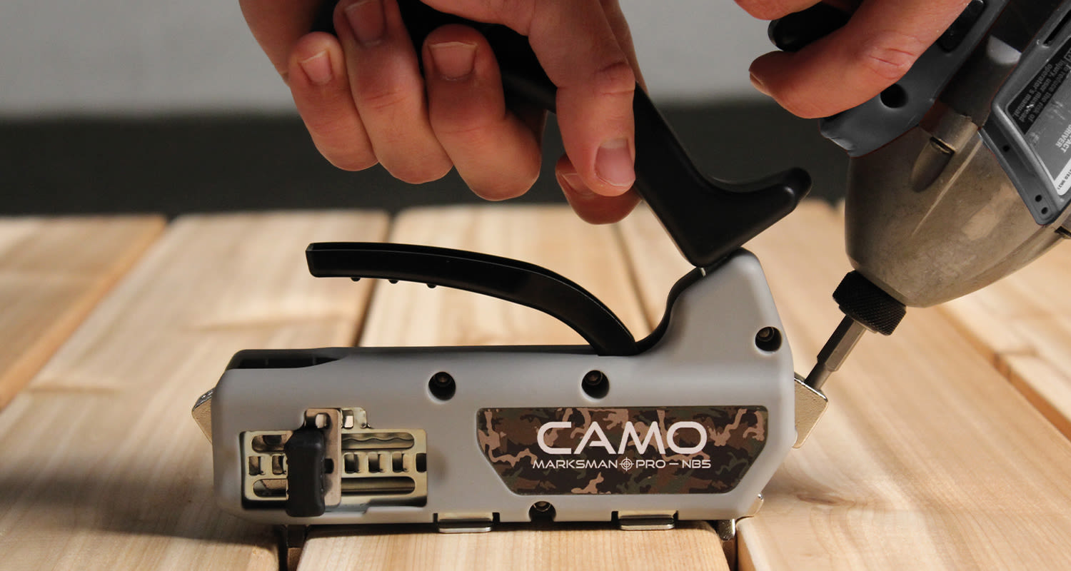 Kaksi kättä pitää harmaata CAMO Marksman PRO -asennustyökalua vaalean puulankun reunalla.