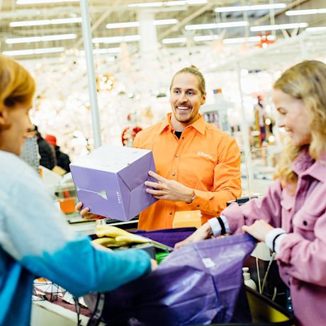 Monivärisessä rautakaupassa oranssipaitainen kassa ojentaa violetin laatikon asiakkaalle.