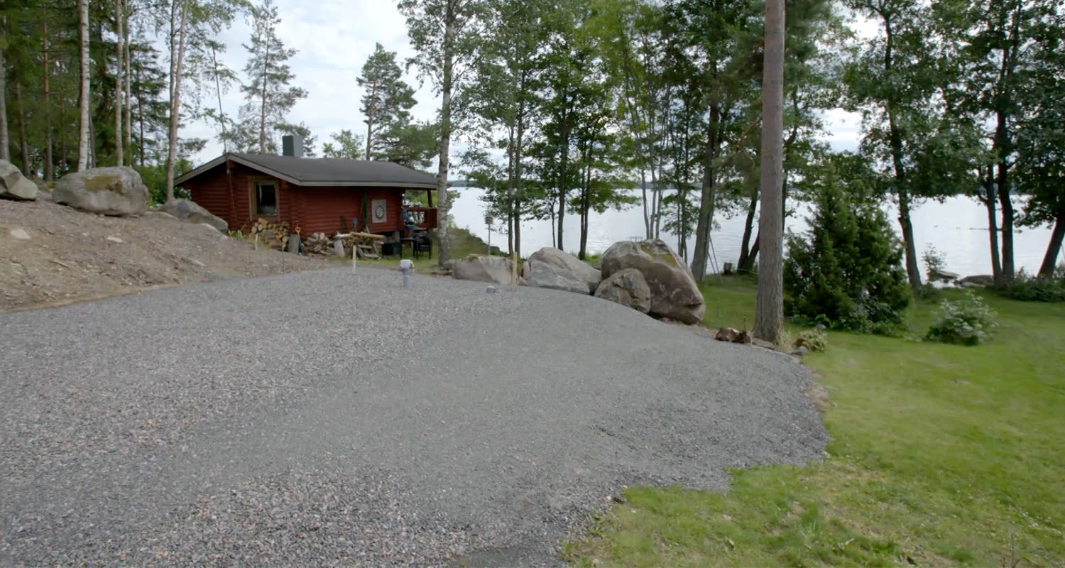 Punainen mökki harmaalla sorapihalla, kivikkoisia rantametsiä ja sininen järvi taustalla