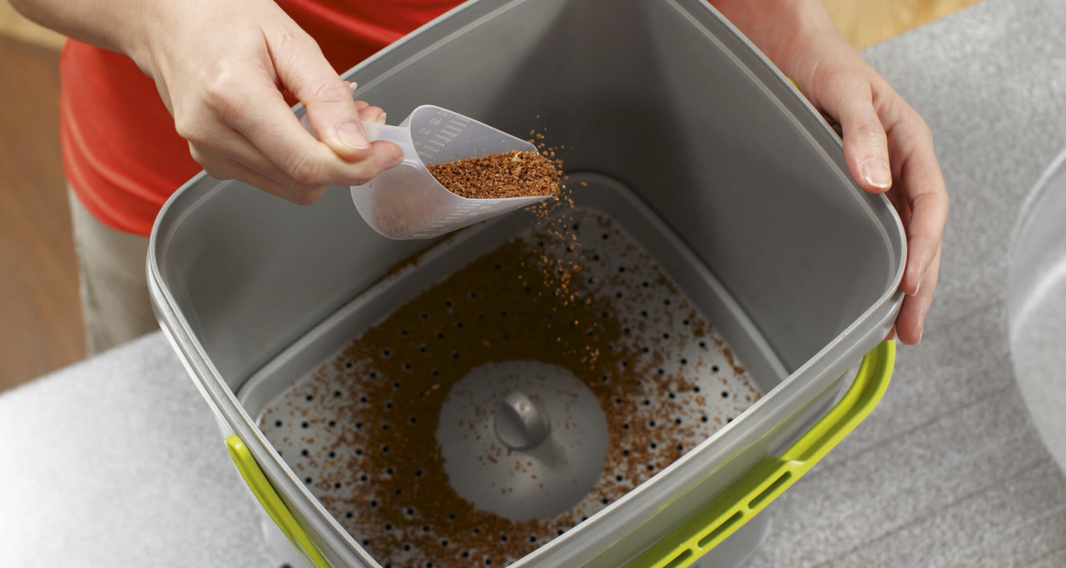 Käsi lusikoi ruskeita kahvinpuruja harmaaseen ämpäriin limenvihreällä kahvalla